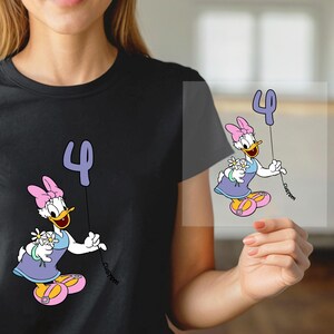 May include: Black t-shirt featuring a cartoon Daisy Duck holding a number 4 balloon and flowers. Daisy Duck is wearing a purple dress, pink bow, and yellow shoes. The word "custom" is written on the balloon string.