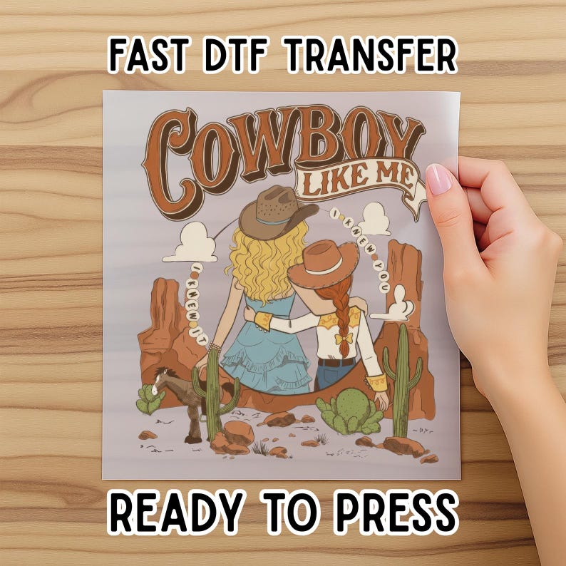 May include: A DTF transfer sheet featuring a graphic illustration of two figures with blonde and red hair wearing cowboy hats, standing in a desert landscape with cacti, rocks, and a horse. The text reads "COWBOY LIKE ME" and "I KNEW YOU".