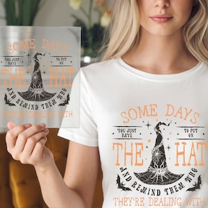 May include: A white t-shirt and a clear square sign with the text "Some Days You Just Have To Put On The Hat And Remind Them Who They're Dealing With" in orange and black. The design features a witch's hat and spiderweb.