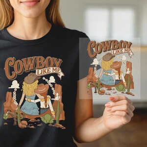 May include: A black t-shirt featuring a graphic illustration of two people wearing cowboy hats in a desert landscape with cacti and rock formations. The text "COWBOY LIKE ME" is printed in a brown, stylized font above the figures.