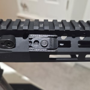 May include: Close-up of a black metal firearm rail system with a series of numbered slots and a small, textured attachment. The attachment has the text "#NANO" and a circular symbol. The rail is designed for accessories.