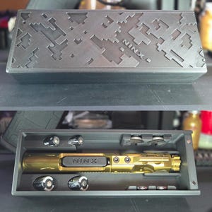 May include: Black rectangular case with a pixelated design on the lid. Inside, a gold-colored object with the text "NINX" is visible, along with batteries and small metal discs. The case is open, revealing its contents.