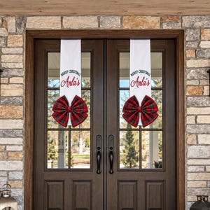 May include: Two white vertical door banners hanging on double glass doors. Each banner features the text Merry Christmas in green and the name Anla's in red script, finished with a large red and green plaid fabric bow at the bottom.