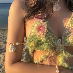 May include: A seashell-themed beachwear set. The set includes a floral bikini top with pink and green accents, a seashell necklace, and a seashell arm cuff. The overall aesthetic is soft and feminine, with a focus on natural elements.