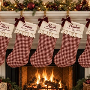 Puede incluir: Cinco calcetines de Navidad con estampado de cuadros vichy rojo y blanco, un puño color crema con borde de volantes y un lazo de terciopelo rojo oscuro. Cada calcetín está bordado con un nombre y el texto Baby's First Christmas 2026.