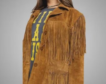 May include: A tan suede jacket featuring long fringe detailing across the chest, shoulders, and pockets. The jacket has a collared neckline, a button-front closure, and is worn over a dark gray t-shirt with yellow block lettering.