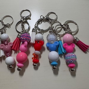 May include: Five keychains featuring colorful beads, including spherical shapes, mouse-ear silhouettes, and dangling tassels. The beads are in shades of pink, white, red, and blue, with some featuring glittery textures. Each keychain has a metal ring.