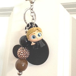 May include: A keychain featuring a cartoon character with blonde hair and blue eyes, wearing a black outfit. The keychain includes black and brown beads, and a silver-colored clasp.