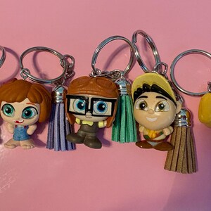 May include: A collection of keychains featuring characters from the animated film "Up." Each keychain has a silver ring, a tassel, and a miniature figurine of a character, including Carl, Ellie, Russell, and Dug.