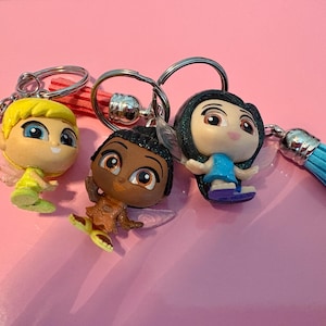 May include: Three fairy-themed keychains with silver key rings and colorful tassels. The figurines have large eyes and are wearing dresses in yellow, brown, and blue. The background is a solid pink color.