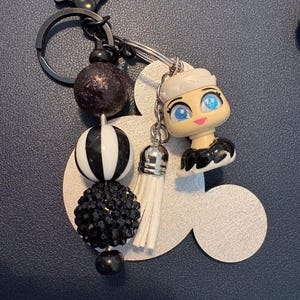 May include: A decorative keychain featuring a black and white beaded design, a small doll figure, and a silver tassel. The keychain includes a black metal ring and a Mickey Mouse-shaped charm. The beads are in various shapes and sizes, with a mix of textures.