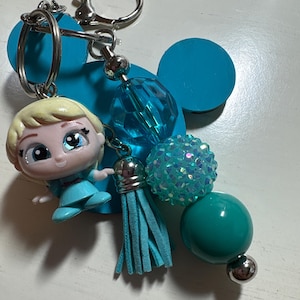 May include: A keychain featuring a light blue silhouette of a mouse head, a small blonde character figurine, a light blue suede tassel, and a series of beads in shades of teal, turquoise, and iridescent mint green, all attached to a silver metal ring.