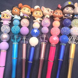 May include: A collection of colorful pens with decorative toppers featuring cartoon characters and beaded accents. The pens have various colors, including blue, pink, and gold, and are arranged in a row. The toppers include a variety of characters and decorative beads.