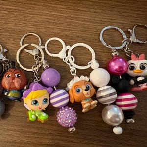 May include: A collection of keychains with character figurines and decorative beads. The keychains feature various characters, including a dog, a duck, and other cartoon figures. The beads are in various colors, including purple, pink, and silver.