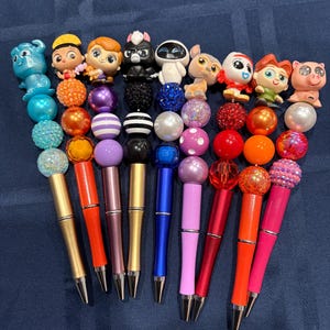 May include: A collection of nine pens, each topped with a unique character figurine and a series of colorful, textured beads. The pens feature various metallic and solid-colored barrels, including gold, orange, purple, blue, pink, and red shades.