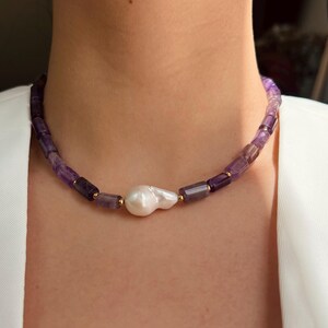 May include: A necklace featuring amethyst beads and a large, irregular white pearl. The amethyst beads are a mix of purple hues, and the necklace has gold-colored accents. The necklace is a jewelry item.