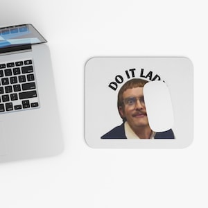 May include: White rectangular mousepad with the text "DO IT LAD" above a man with glasses and a mustache. A white computer mouse is on the mousepad. A laptop is in the background.