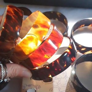May include: A collection of tortoiseshell-patterned bangles in shades of brown, amber, and red. The bracelets are stacked together, showcasing their glossy finish and unique color variations. The image captures the light reflecting off the smooth surfaces of the jewelry.