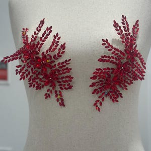 May include: Two red rhinestone floral appliques, each with a cluster of flowers and leaf-like details. The appliques are displayed on a mannequin, suggesting their use in fashion or craft projects. The rhinestones sparkle, adding a touch of glamour to the design.