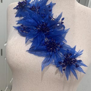 May include: A royal blue floral applique featuring layered sheer fabric petals and dark blue crystal bead embellishments. The decorative piece is arranged in a cluster of three distinct flower shapes with thin wire stems.