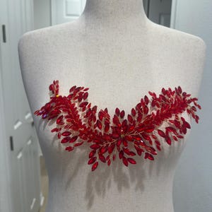 May include: A red crystal and gold wire bridal headpiece, shaped in a curved design. The headpiece is displayed on a mannequin. The crystals are a deep red colour, and the wire is gold. The design is reminiscent of a floral arrangement.