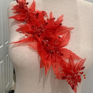 May include: A vibrant red floral applique, featuring layered petals and beaded accents. The design is attached to a mannequin, showcasing the intricate details of the fabric and embellishments. The applique is a striking decorative element.