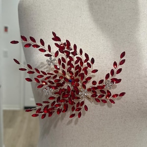 Può includere: Un accessorio per capelli rosso e argento con un design floreale. Presenta elementi a forma di foglie di cristallo rosse e dettagli argentati, con una piccola gemma blu al centro. Esposto su un manichino.