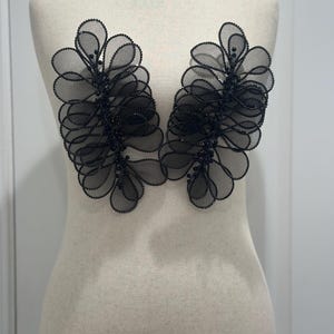 May include: Two black floral appliques with beaded accents. The appliques are made of layered, sheer fabric, creating a three-dimensional effect. The design is symmetrical, with each applique resembling a stylized flower or leaf arrangement. They are displayed on a mannequin.