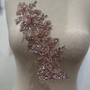 May include: A floral applique with clear and pink crystals arranged in a cascading design. The embellishment features a variety of crystal shapes, including teardrops and faceted stones, creating a sparkling effect. The applique is likely for bridal or formal wear.