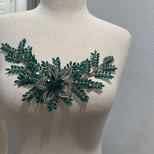 May include: A decorative emerald green and silver applique, shaped like a floral design, is displayed on a mannequin. The applique features numerous green rhinestones and small black beads, creating a sparkling, ornate embellishment.