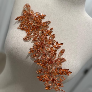 May include: An orange and gold floral applique with sparkling rhinestones. The embellishment is arranged in a cascading design, featuring multiple flower and leaf shapes. The decorative piece is affixed to a mannequin.