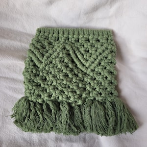 May include: A handmade, green macrame coaster with a geometric pattern and fringe detail. The coaster is square-shaped and made of thick, woven cotton cord. The fringe hangs from the bottom edge.