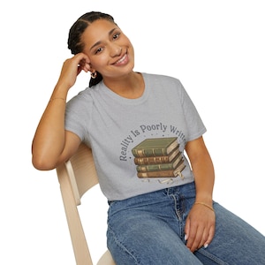 May include: A light grey t-shirt with a graphic of a stack of books and the text "Reality Is Poorly Written." The books are in shades of green and brown, with a white ribbon bookmark. The shirt is worn with blue jeans.