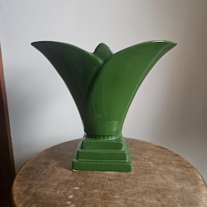 May include: A green ceramic vase with a tulip-like shape. The vase has a square base and a flared top with pointed edges. The vase is a solid, glossy green color and sits on a wooden surface.