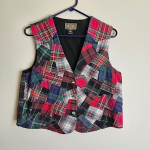 May include: A patchwork vest with a V-neck design. The vest features a variety of plaid patterns in red, green, blue, and black. It has gold-colored buttons and a black lining. The vest is hanging on a blue hanger.