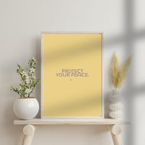 May include: A framed print with the text "PROTECT YOUR PEACE" in purple on a yellow background. The print is displayed on a light-coloured wooden shelf, with a white vase holding flowers on the left and a white vase with dried grass on the right.