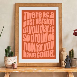 May include: A framed art print with the text "There is a past version of you that is so proud of how far you have come." The text is in a coral color against an orange background. The frame is made of wood.