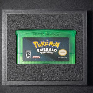 以下が含まれることがあります： ゲームボーイアドバンス用ゲームソフト「ポケットモンスターエメラルドバージョン」のフレーム入りカートリッジ。カートリッジは緑色で、ゲームのロゴが黄色と青色で描かれた黒いラベルが付いています。フレームは濃いグレーで、黒い背景に配置されています。