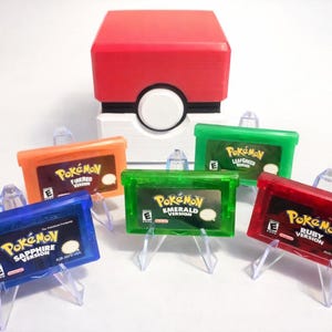 Könnte beinhalten: Ein roter und weißer Pokéball-förmiger Behälter mit fünf farbigen Pokémon-Spielkassetten. Die Kassetten sind orange, grün, rot, blau und grün, jede mit einem anderen Spieltitel versehen. Die Spiele werden auf transparenten Kunststoffständern präsentiert.