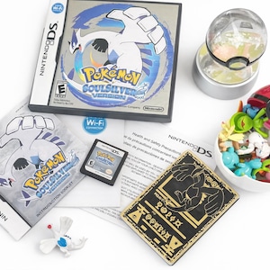 Op de afbeelding: Een verzameling Pokémon-merchandise, waaronder een Nintendo DS-gamecase, gamecartridge, instructieboekje en een rood-witte Poké Ball gevuld met kleine beeldjes. Een doorzichtige, bolvormige container en een goudkleurige kaart zijn ook aanwezig.