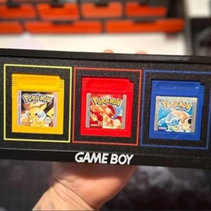 Könnte beinhalten: Ein schwarzer, gerahmter Display mit drei Game Boy-Cartridges. Die Cartridges sind gelb, rot und blau, jede mit einem anderen Pokémon-Spiel-Label. Der Rahmen trägt den Schriftzug "GAME BOY" in weißen Buchstaben.