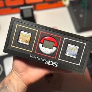 May include: A black rectangular display frame featuring the Nintendo DS logo in white. The frame contains two Nintendo DS game cartridges and a circular red and white Pokéwalker device, each set within individual recessed foam compartments.