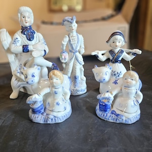 Puede incluir: Colección de figuras de porcelana blanca con detalles azules. Las figuras representan figuras con trajes de época, algunas con animales. La colección incluye una figura sentada, una figura de pie y figuras con vacas. Las figuras tienen adornos dorados.
