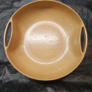 Pode incluir: Uma tigela redonda castanho-clara com duas pegas integradas. A tigela tem uma superfície lisa, ligeiramente texturizada e um design largo e raso. As pegas são curvas e integradas na borda da tigela. A tigela é provavelmente feita de madeira ou material semelhante.