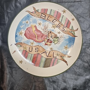 May include: Round decorative plate with a painted design. The plate features a teddy bear angel with wings, a red cloak, and the words "BLESS US ALL" on banners. The background has stars and a blue sky.