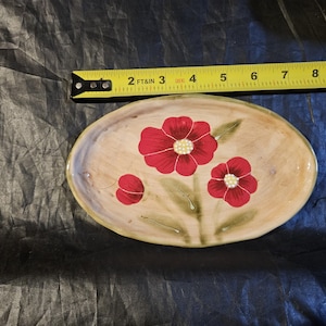 May include: Oval ceramic serving dish with a beige background and red floral design. The dish is approximately 9 inches long and 6 inches wide. The flowers have white centers and green leaves.