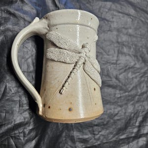 May include: A ceramic mug with a light beige, speckled glaze and a smooth finish. The exterior features a prominent, raised relief sculpture of a dragonfly with textured wings and a segmented body. It has a curved handle and a cylindrical shape.