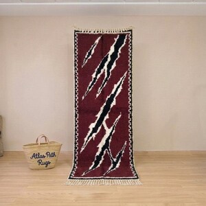 May include: A long, rectangular rug with a burgundy background and a pattern of black and white diagonal slashes. The rug has a fringed edge and is displayed against a neutral wall. A straw basket with the text "Atlas Path Rugs" is in the foreground.