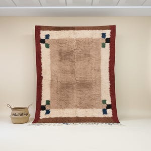 Puede incluir: Una alfombra rectangular grande con un campo central marrón, bordeada por crema y rojo oscuro. Las esquinas presentan patrones cuadrados de negro, verde y azul. Una cesta tejida con el texto "Atlas Path Rugs" está cerca.