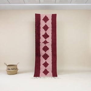May include: A long, rectangular rug featuring a central light pink diamond pattern framed by deep burgundy. The rug has a textured, plush appearance and a fringe at the bottom. A woven basket is in the lower left corner.